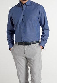 Chemise à carreaux bleue et blanche à boutons avec manches longues, associée à un pantalon de costume gris clair et une ceinture noire.