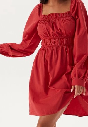 Femme portant une robe rouge à manches longues, col carré, taille et poignets froncés, soulevant légèrement la jupe d'une main.