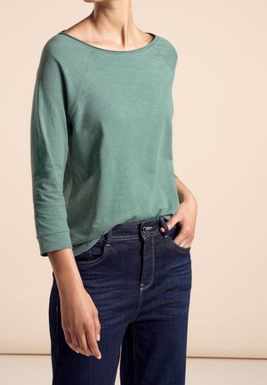 Mujer con una blusa de manga larga verde claro y jeans azul marino, con una mano en el bolsillo, sobre un fondo neutro.