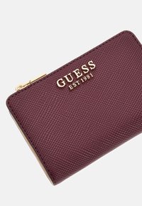 Cartera de cuero texturizado burdeos con el logo dorado de GUESS y la marca EST 1981, que presenta un cierre de broche dorado.