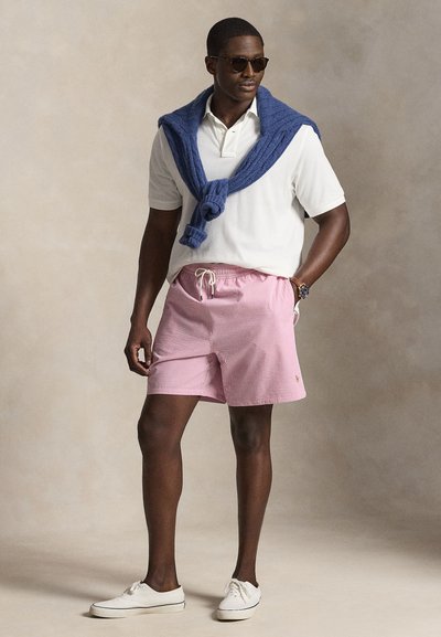 Άνδρας με γυαλιά ηλίου, άσπρο πολο πουκάμισο, ρόζ ριγέ σορτς, άσπρα αθλητικά παπούτσια, με μπλε πουλόβερ που αιωρείται στους ώμους, σε ουδέτερο φόντο.