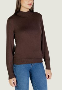 Brązowy sweter dzianinowy z wysokim kołnierzem, prążkowanymi mankietami i pasem. Połączony z niebieskimi dżinsami, prezentującymi dopasowany, casualowy styl.