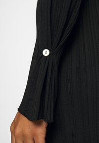 Manche en tissu côtelé noir avec un détail plissé et une fermeture bouton circulaire. La texture est douce avec une légère élasticité.