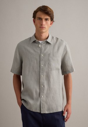 Junger Mann mit braunen Haaren, der ein hellgraues, kurzärmeliges Hemd mit Knöpfen und dunkle Hosen vor einem schlichten Hintergrund trägt.