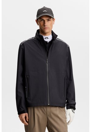 Schwarze wasserdichte Jacke mit durchgehendem Reißverschluss, Seitentaschen und elastischen Bündchen. Dazu wird ein weißes Hemd mit Kragen und eine passende Mütze mit Logo getragen.