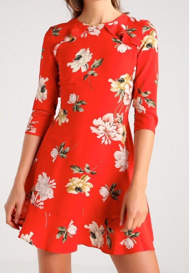 Femme portant une robe rouge mi-longue ornée de motifs floraux blancs et jaunes, avec des manches trois-quarts, debout devant un fond uni.