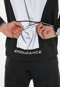 Sort og hvid atletisk top med reflekterende detaljer. Har en lynlåslomme på bagsiden. "ENDURANCE" logo på den nederste kant.