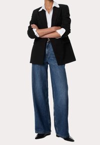 Sort dobbeltradet blazer over hvid skjorte med krave, parret med brede blå jeans og spidse sorte sko. Rette linjer, struktureret pasform.