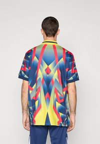 Tričko s krátkým rukávem v modré, žluté a červené barvě. Obsahuje geometrické vzory s přechodovým efektem. Doplňuje ho žlutý límec a modré manžety.