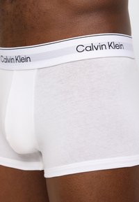 Κοντινό πλάνο ενός ατόμου που φοράει λευκά μποξεράκια Calvin Klein με ζώνη μέσης με το λογότυπο, σε σκούρο τόνο δέρματος.