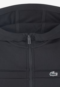 Must värviline lukuga tunkedega pusa, millel on sile kangatekstuur, lamedad õmblused ja väike vasakul rinnal tikitud logo.