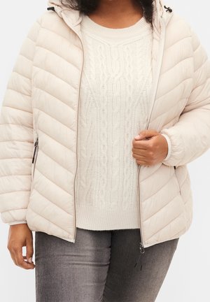 Personne portant un pull en maille torsadée crème sous une veste matelassée beige clair assortie à un jean gris, visible du cou aux hanches.