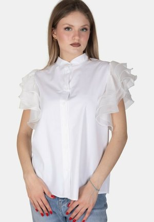 Giovane donna che indossa una camicetta bianca con maniche a volant sovrapposti e jeans azzurri, in piedi davanti a uno sfondo uniforme.