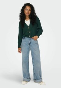 Cardigan verde a coste con grandi bottoni, indossato sopra una maglietta bianca. Abbinato a jeans azzurri a gamba larga e sneakers bianche.