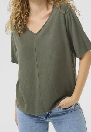 Frau mit welligem blondem Haar, die ein olivgrünes V-Ausschnitt-Kurzarmshirt und hellblaue Jeans trägt, mit der Hand in der Tasche.