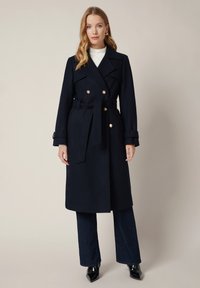 Granatowy wełniany płaszcz trench z podwójnymi złotymi guzikami, z paskiem w talii i szerokimi klapami, w zestawieniu z ciemnymi dżinsami i czarnymi szpilkami.