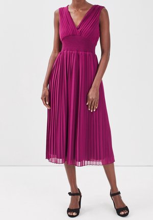 Robe midi plissée sans manches d'un rose profond avec une taille cintrée, décolleté en V, portée avec des talons noirs à bride à la cheville sur un mannequin.