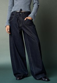 Tmavomodré rifľové nohavice s širokými nohavicami a sťahovacou šnúrkou v páse, zdobené kontrastným prešívaním a bočnými vreckami, v kombinácii s rebrovaným sivým svetrom.