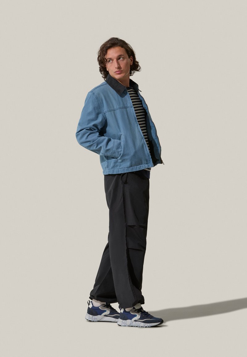Giacca di jeans blu chiaro con colletto nero, indossata sopra una camicia a righe bianco e nera, abbinata a pantaloni neri e sneakers blu.