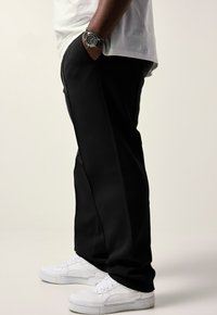 Persoon met zwarte broek, witte sneakers en een wit shirt, met een zwart horloge en armband om de linkerpols, die zijwaarts staat.