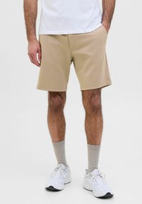 Shorts beige avec une texture lisse, présentant des poches latérales et un ourlet cousu, portés avec des baskets blanches et des chaussettes gris clair.