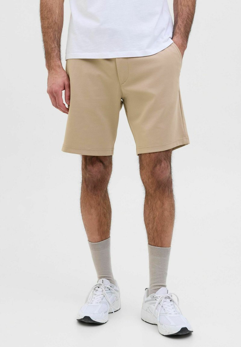 Shorts beige avec une texture lisse, présentant des poches latérales et un ourlet cousu, portés avec des baskets blanches et des chaussettes gris clair.
