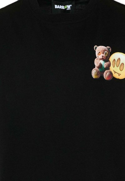 Camiseta negra con un oso de peluche que sostiene un corazón verde y una cara de emoji amarilla con tres ojos y una boca con colmillos en el pecho.