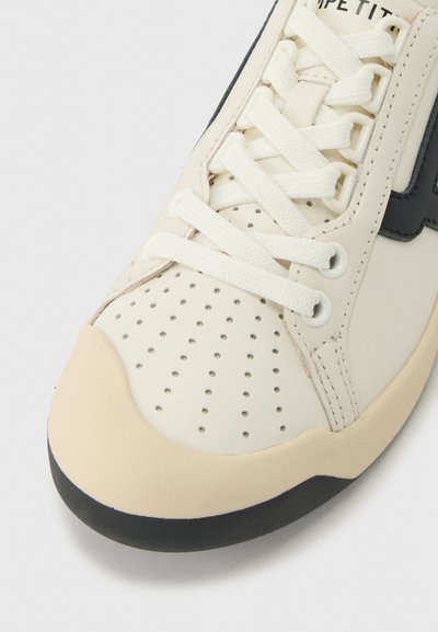 Gros plan sur l'avant d'une chaussure de sport beige avec des perforations, des lacets blancs, un bord de semelle noir et des détails de piqûres visibles sur le dessus.