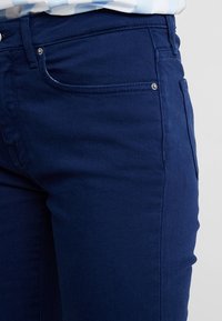 Jeans de mezclilla azul oscuro con un diseño clásico de cinco bolsillos, detalles de costura y una textura suave. Detalles metálicos simples en la parte frontal.