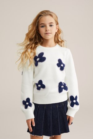 Balts džemperis ar tumši zilām pūkainām ziedu apdrukām, apaļš kakla izgriezums un ribota apakša. Saskaņots ar tumši džinsa plisētu svārku.