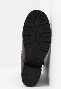 Hnědá kožená bota s robustní černou gumovou podrážkou, která má výrazný vzor a texturované detaily. Logo je vyražené na podrážce.