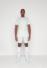 Lys grå sportantrekk med en tettsittende t-skjorte og shorts, begge med en cyan Nike-logo. Hvite knelange sokker og lyseblå sko fullfører settet.