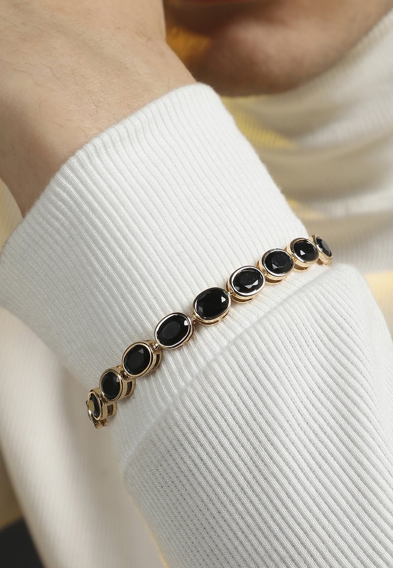 Bracciale oro con pietre nere ovali, incastonate in maglie metalliche, indossato sopra il polsino di un maglione bianco a coste.