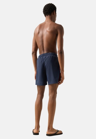 Homme debout pieds nus, de dos, portant un short de bain bleu marine avec une ceinture élastique et des tongs noires sur un fond clair.