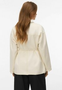 Pikkade laineliste juustega naine, kes kannab helebeeži vööga jope ja musti pükse, seisab seljaga vastu tavalist tausta.