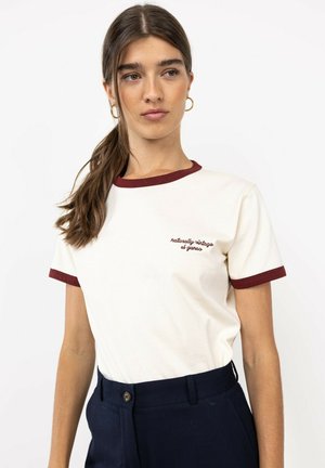 Mujer joven con el pelo largo y castaño recogido en una coleta lleva una camiseta blanca con ribetes granate y pantalones azul marino, mirando ligeramente hacia un lado.