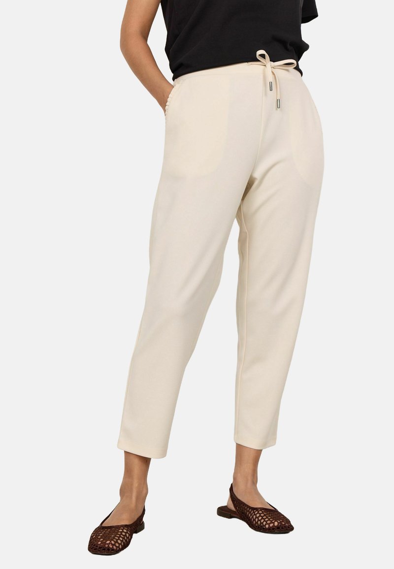 Cream-colored tapered pants with a smooth texture, adjustable drawstring waist, side pockets, and a relaxed fit, paired with brown flat shoes.
