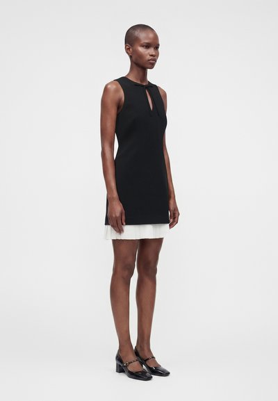Femme portant une robe noire sans manches avec une ouverture clé à l'avant et un ourlet plissé blanc, associée à des escarpins noirs Mary Jane, debout devant un fond uni.