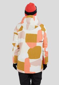 Personne portant une veste à capuche avec des blocs de couleur abstraits rose, blanc et moutarde, un bonnet rouge, des gants noirs et un pantalon noir, de dos.