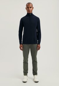 Maglione blu navy con collo alto e zip a quarto, abbinato a pantaloni verde oliva e sneakers bianche. Design semplice, silhouette aderente.