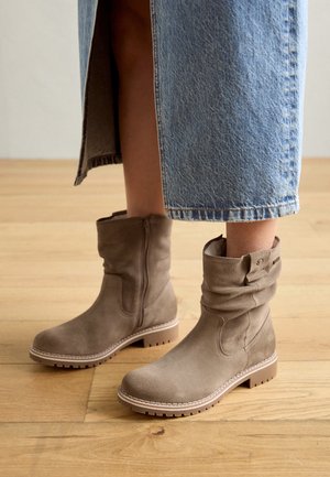 Bottes mi-mollet en daim marron clair avec fermeture éclair latérale et détail de bride portées avec une longue jupe en jean bleu sur un sol en bois.