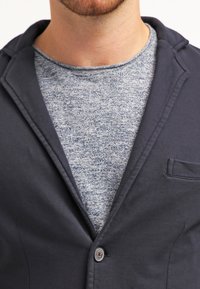 Blazer navy scuro con rever a punta e chiusura con un bottone, indossato sopra una maglia grigia a girocollo con texture. Dettaglio della tasca visibile sul blazer.
