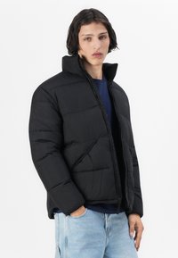 Sort pufferjakke med høj krave, lynlås foran og quiltet design. Tekstureret stof og afslappet pasform, parret med lyseblå jeans.