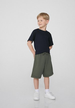 Marineblauw katoenen T-shirt gecombineerd met olijfgroene shorts met een trekkoord in de taille en een opgerolde zoom. Draagt witte sportschoenen.