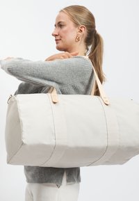 Borsa da viaggio in tessuto beige con due manici in pelle, forma cilindrica ampia e dettagli cuciti. Donna in maglione grigio la tiene.