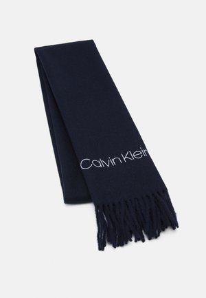 Bufanda azul oscuro doblada con extremos con flecos y el logo "Calvin Klein" bordado en blanco a lo largo de un borde.