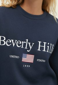 Felpa blu marina con testo bianco "Beverly Hills", piccola bandiera degli Stati Uniti, testo "UNITED STATES" e "1999" sotto la bandiera.
