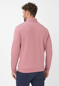 Uomo con capelli ricci corti che indossa un pullover a maniche lunghe di un rosa pallido e pantaloni blu navy, volto di spalle su uno sfondo bianco uniforme.
