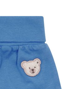 Niebieska tkanina z haftowaną białą twarzą misia w pobliżu pasa, ukazująca szczegółowe szwy i fakturę dzianiny.