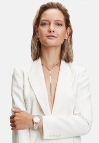 Silberne Lasso-Halskette mit einem geschwungenen Design. Das Modell trägt einen weißen Blazer und eine silberne Uhr mit einem verlinkten Armband. Der Hautton ist neutral.